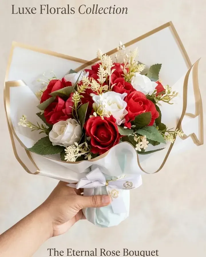 Rosas artificiais vermelhas e brancas com folhagens e embalagem clara com detalhes dourados.
